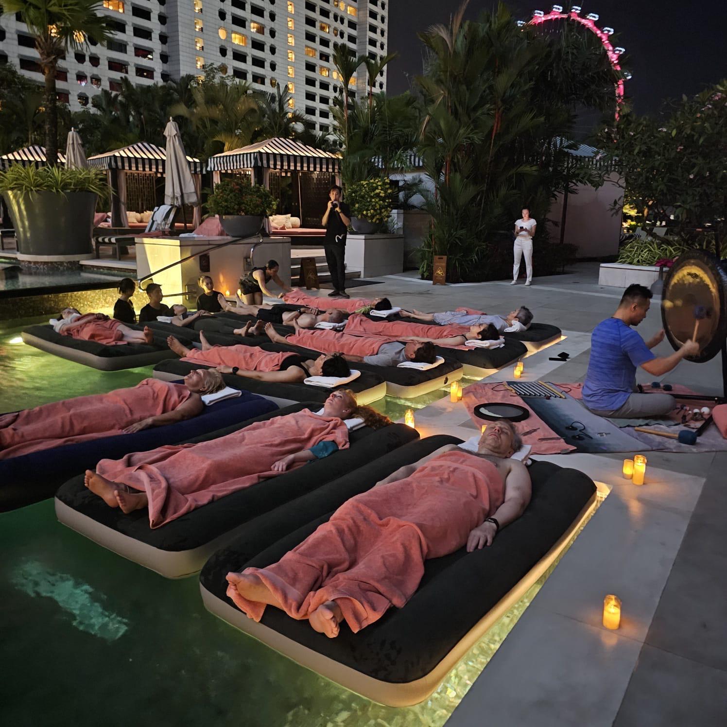 Participants resting on floating mattresses during an outdoor poolside Sound Bath at night